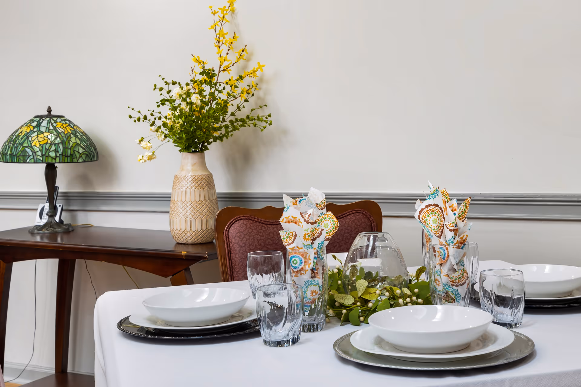 Dining table set with white plates, glasses, patterned napkins and a greenery centerpiece, with a sideboard, lamp, and vase behind it.