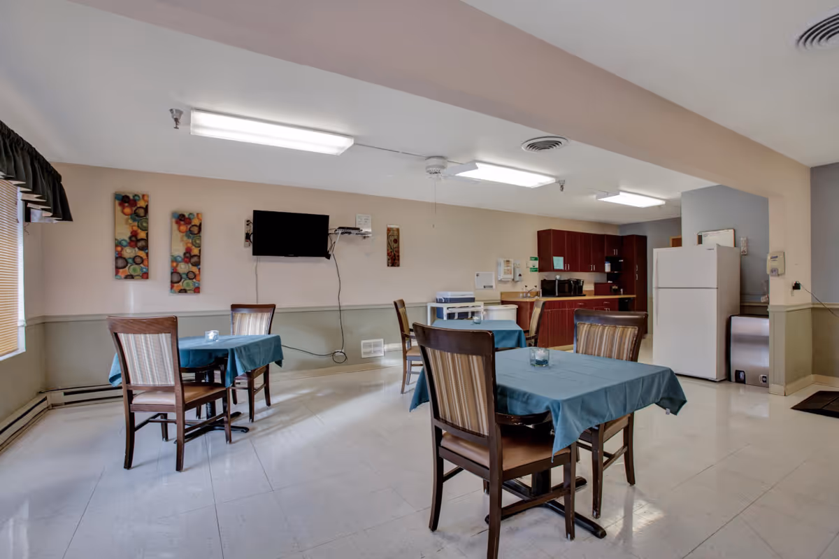 A small dining area with several tables covered with blue tablecloths and wooden chairs. The room has a kitchen area with a white refrigerator, wooden cabinets, a microwave, and a coffee maker. A flat-screen TV is mounted on the wall, and there are colorful abstract wall decorations. The room is well-lit with fluorescent ceiling lights and has a window with blinds and a dark valance.
