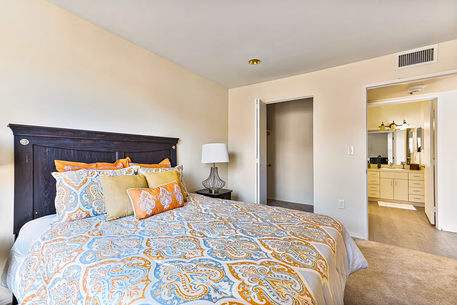 Well-lit bedroom with a patterned bed and dark wood headboard, a bedside lamp, and an open doorway revealing a bathroom and closet.