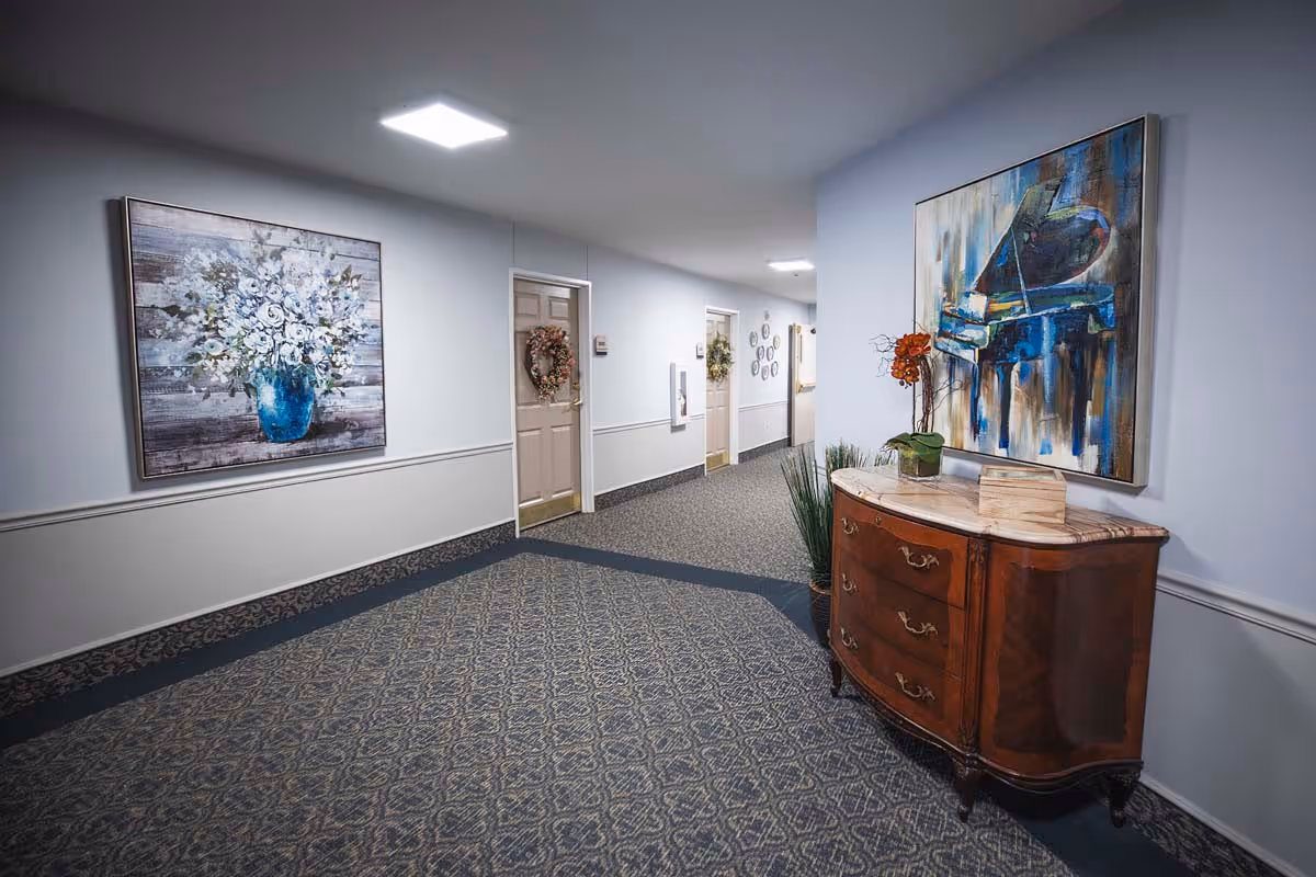 Interior hallway of a senior living facility with patterned carpet, light blue walls, and several doors decorated with wreaths. There is a wooden chest of drawers with a marble top on the right side, adorned with a small plant and a decorative item. Two large paintings hang on the walls, one depicting a bouquet of flowers and the other a piano.