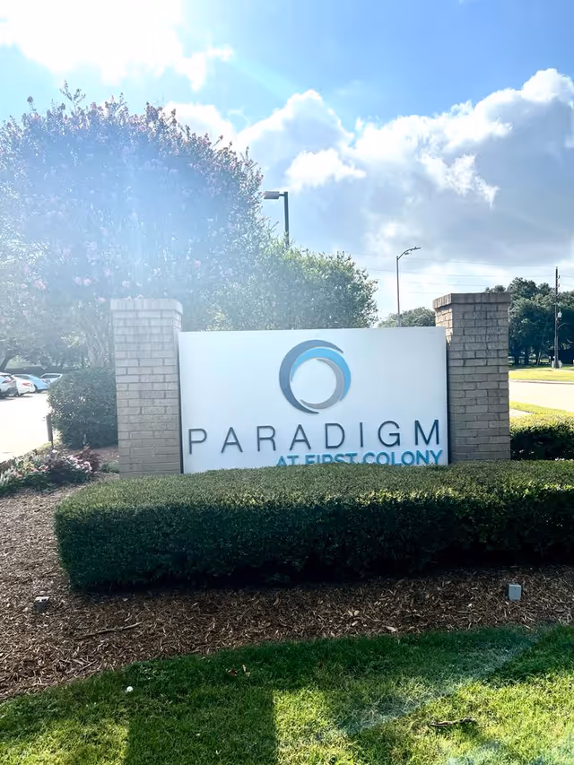 Outdoor view of a sign for Paradigm at First Colony, set between two brick pillars with neatly trimmed bushes and trees in the background under a partly cloudy sky.