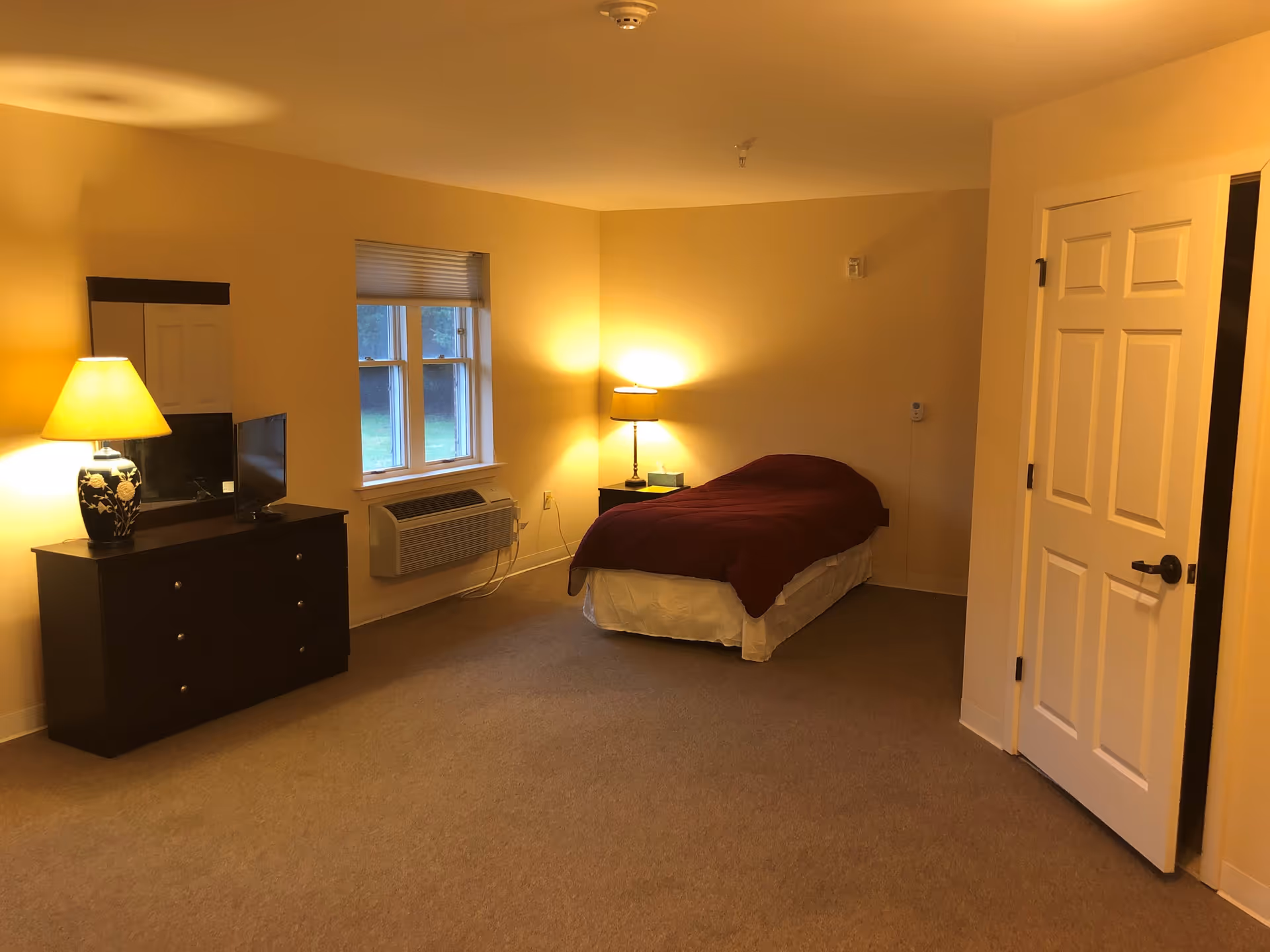 A simple bedroom with beige walls and carpet, featuring a single bed with a maroon blanket, a nightstand with a lamp, a dresser with a lamp and a small TV, and a window with blinds. A white door is partially open on the right side of the room.
