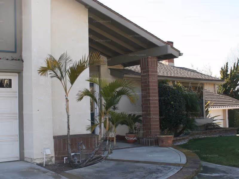 Exterior view of a residential-style building with a covered porch supported by a brick column. There are several potted plants and palm trees near the entrance, a curved concrete walkway, and a well-maintained lawn area. The building has a light-colored stucco exterior and a tiled roof.