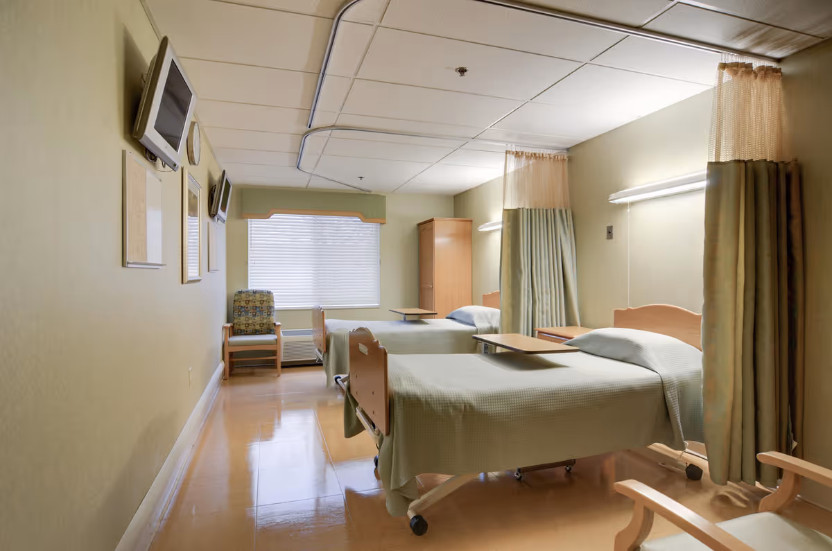 A clean and well-lit room with two hospital-style beds separated by privacy curtains. Each bed has a small overbed table. There is a window with blinds at the far end, a wooden chair with patterned upholstery, a wooden cabinet, and two wall-mounted televisions on the left wall.