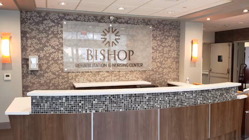 Reception desk with a curved tiled counter and a wall sign reading "BISHOP Rehabilitation & Nursing Center" in a facility lobby.
