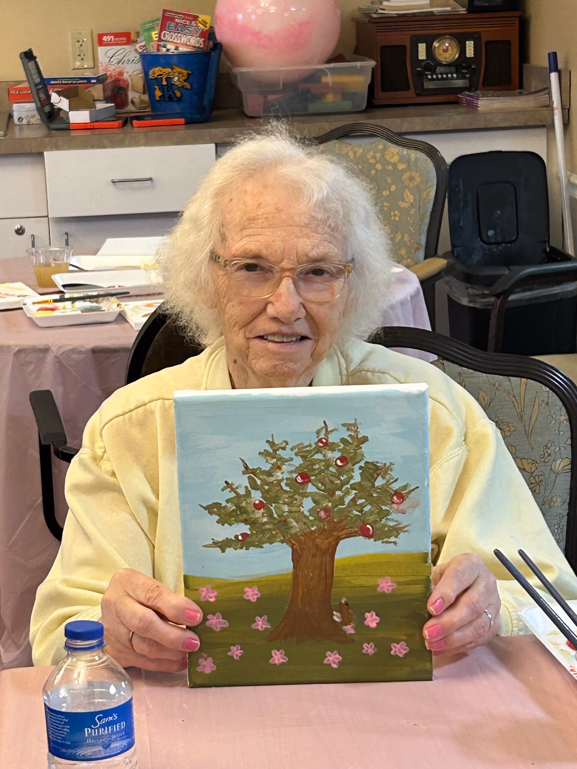 An elderly woman with white hair and glasses is sitting at a table in a room, holding up a painting of a tree with red apples and pink flowers on the ground. The table has art supplies and a water bottle on it. Behind her are chairs, a countertop with various items, and a vintage radio.