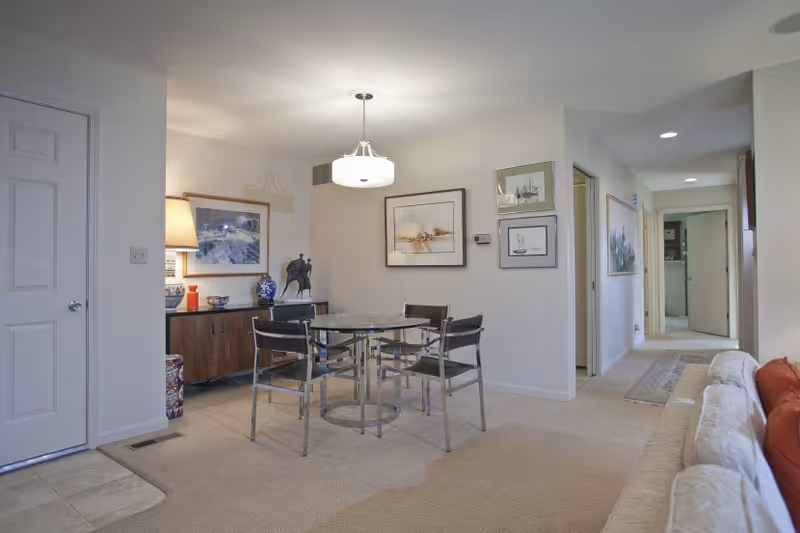 Interior view of a retirement community apartment showing a dining area with a round glass table and four chairs. The room has beige carpet and white walls adorned with framed artwork. A sideboard with decorative items and a lamp is visible in the corner. To the right, a hallway leads to other rooms, and part of a beige sofa with red cushions is visible in the foreground.