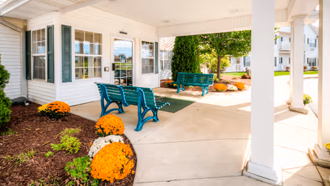Covered outdoor seating area with green benches, surrounded by flower beds with orange flowers and a white building with large windows in the background.