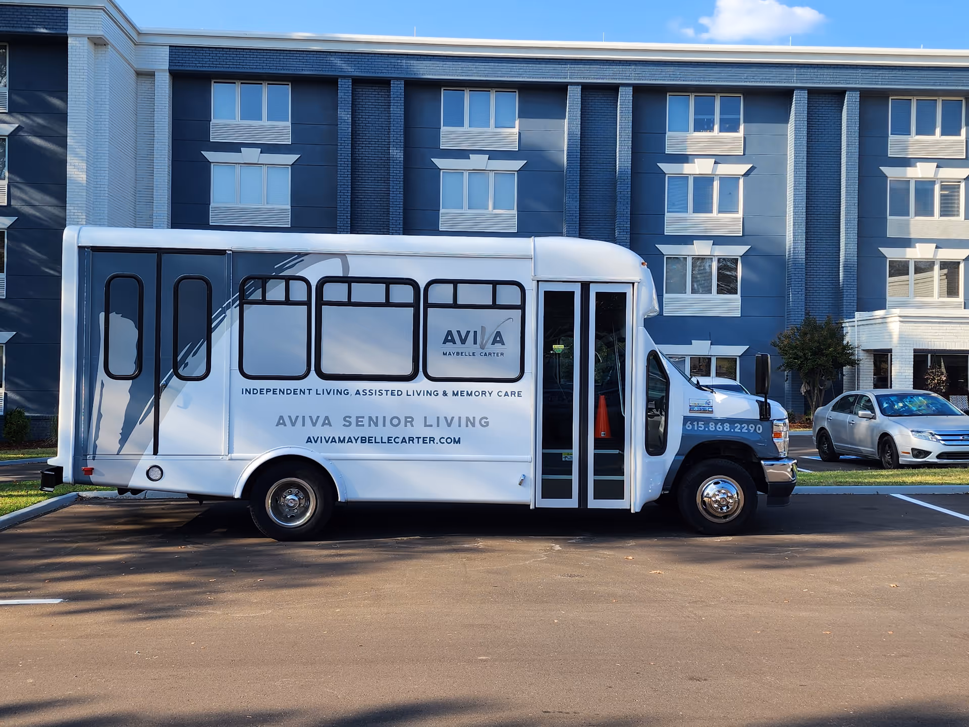 A white shuttle bus parked in front of a multi-story senior living facility building. The bus has the logo and text for AVIVA Maybelle Carter, advertising independent living, assisted living, and memory care services. The building behind the bus is blue and white with multiple windows.