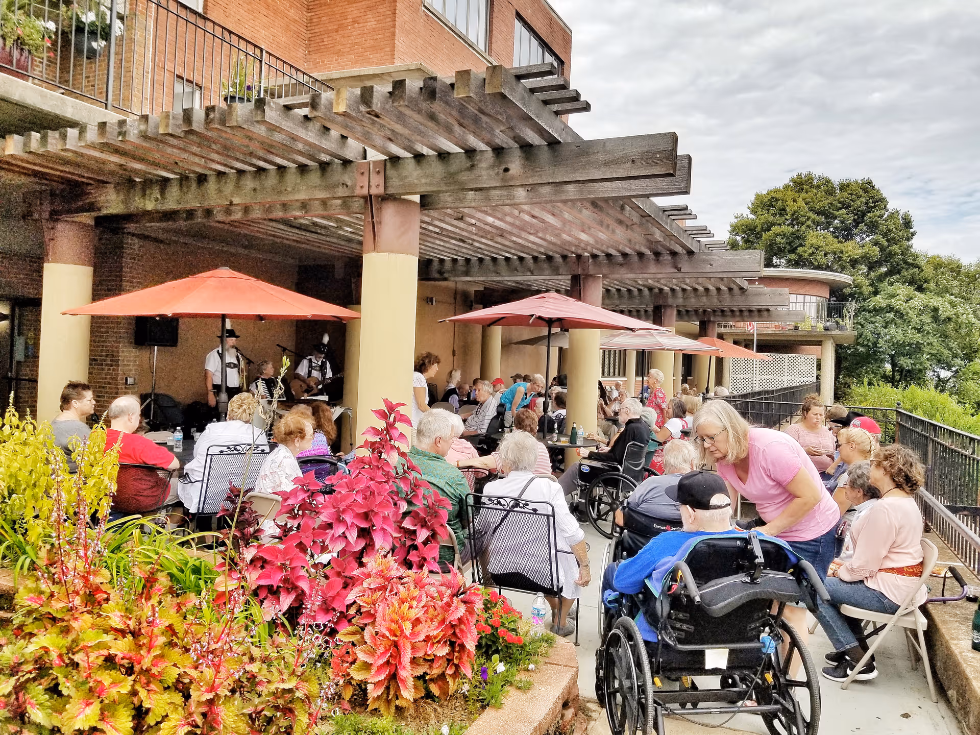 People gathered on a shaded outdoor patio at a senior living facility, sitting at tables under umbrellas with plants in the foreground.