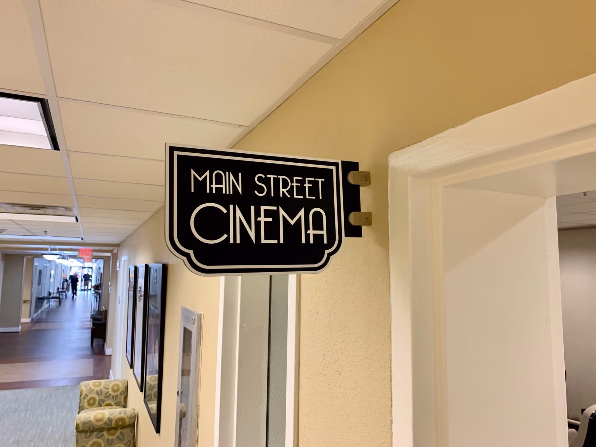 A hallway in a senior living facility with a black and white sign reading 'Main Street Cinema' mounted on a beige wall above a doorway. The hallway has framed pictures on the walls, a patterned armchair, and a person walking in the distance.