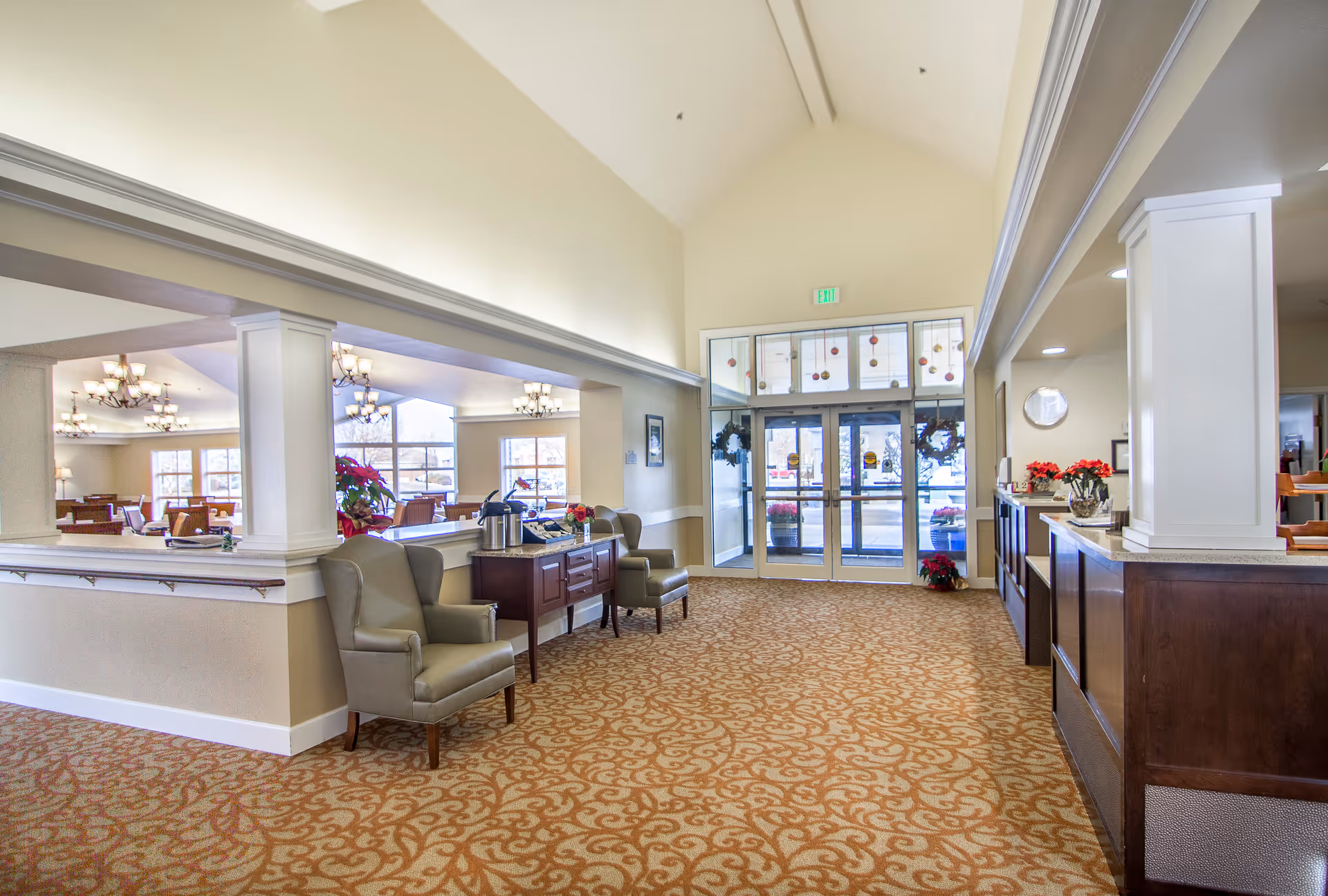 Spacious and well-lit interior lobby area of Brookdale Boise Parkcenter with patterned carpet, two armchairs, a wooden side table with a coffee dispenser, and a reception desk on the right. Large windows and glass doors at the far end let in natural light, and holiday decorations are visible on the doors and side table.