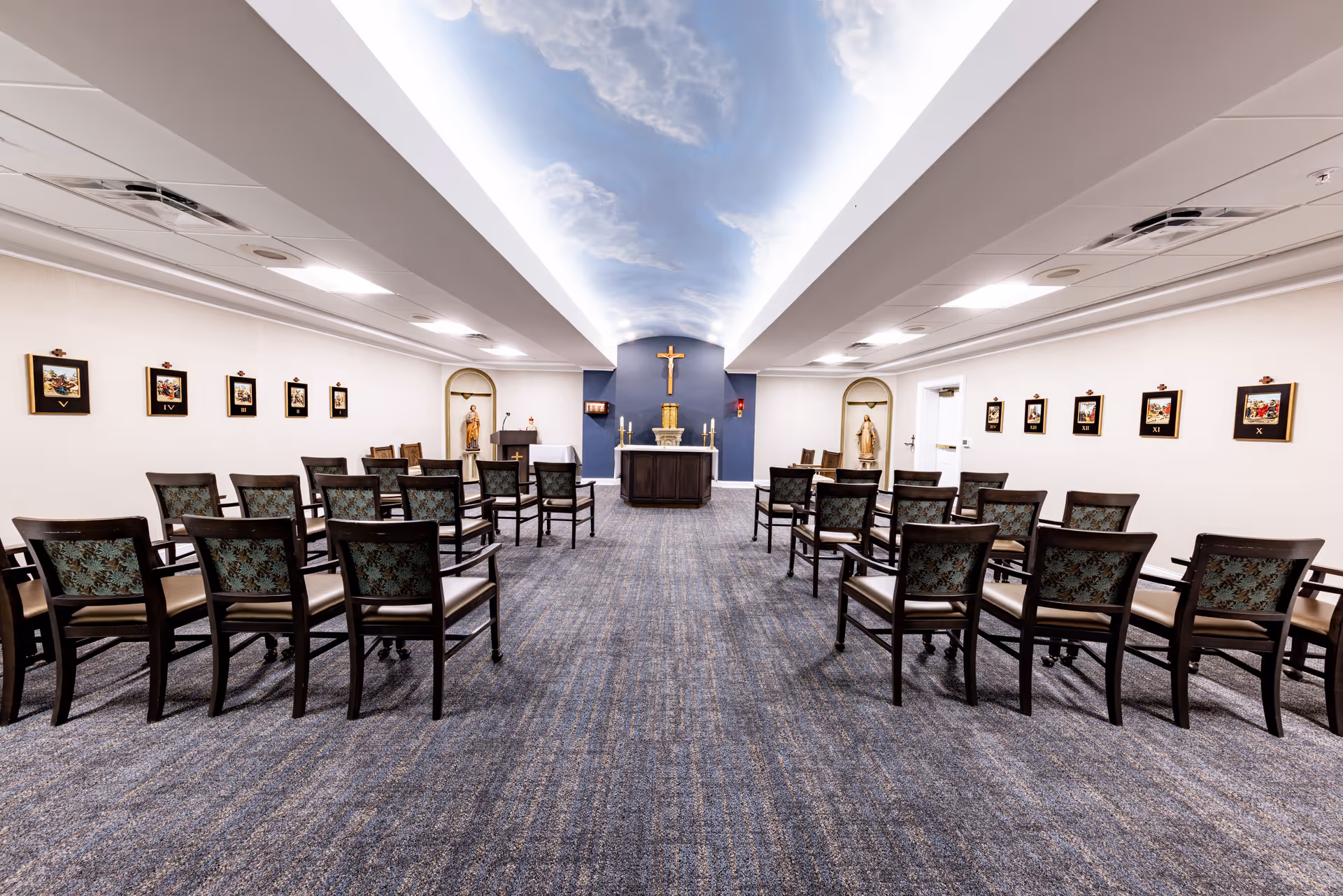Interior view of a chapel or prayer room with rows of wooden chairs facing an altar with a crucifix on the wall. The ceiling features a sky mural with clouds, and religious statues and framed religious images are displayed on the walls.