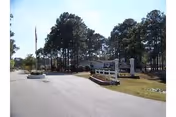 Entrance road leading to a facility with tall pine trees lining the sides and a sign on the right side near the road. The sky is clear and the area is well-maintained with grass and landscaping.