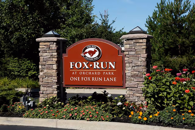 Entrance sign for Fox Run at Orchard Park mounted between stone pillars with flowers and trees around it.