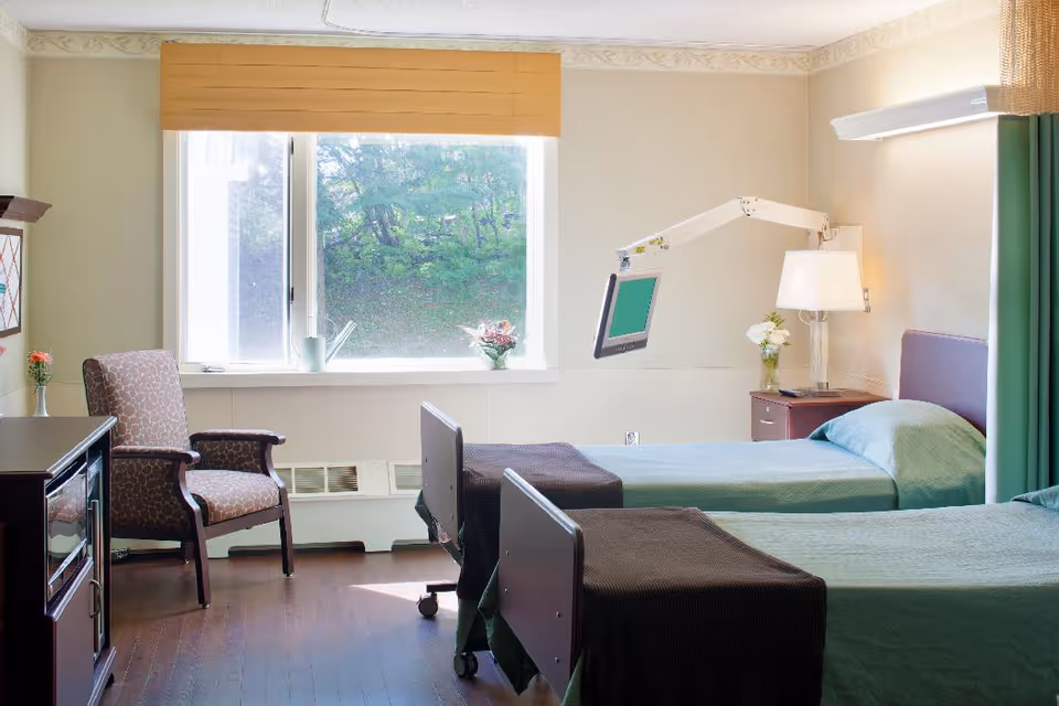 Two single beds in a bright senior-living bedroom with a window, bedside table, chair, and an adjustable TV arm.