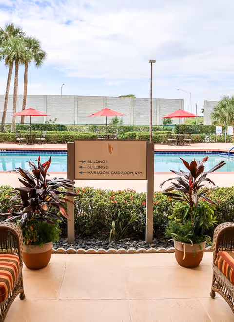 View of an outdoor pool area with red umbrellas and tables in the background, seen from a covered patio with two potted plants and wicker chairs with striped cushions. A sign in the center points to Building 1, Building 2, and Hair Salon, Card Room, Gym.