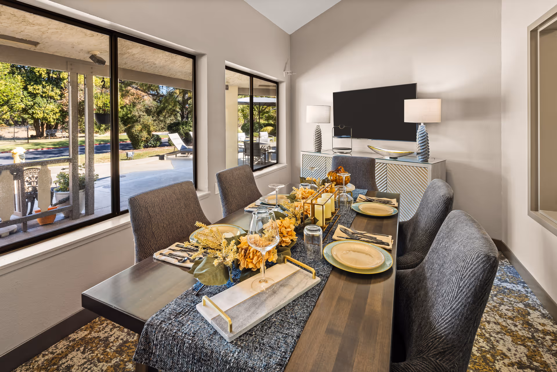 A dining room with a wooden table set for six people, featuring plates, glasses, napkins, and a centerpiece with candles and flowers. The room has large windows showing an outdoor patio area with lounge chairs and greenery. A flat-screen TV is mounted on the wall above a sideboard with two table lamps.