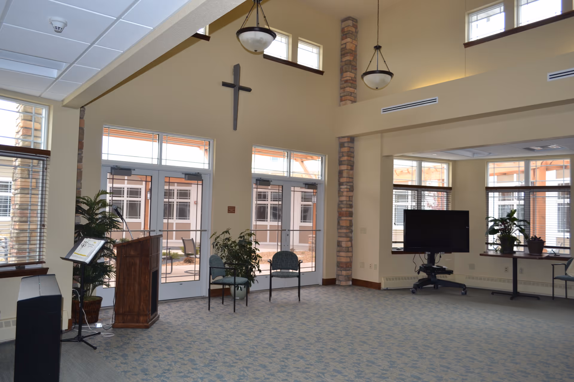 Bright, high-ceilinged common room with large windows and doors, chairs, a podium, a TV on a stand, and a cross mounted on the wall.