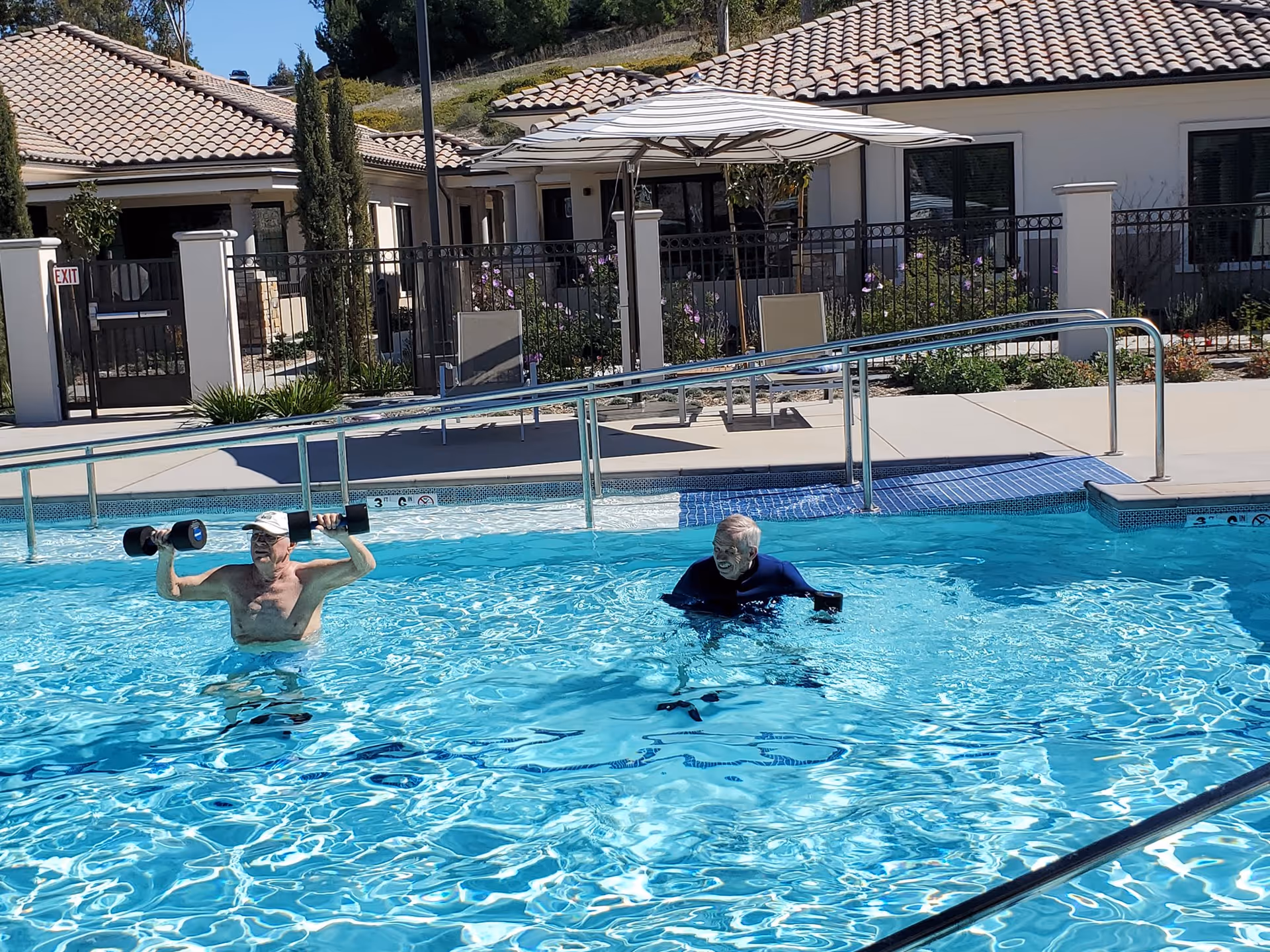 Two elderly men exercising in a swimming pool at a senior living facility. One man is lifting water dumbbells above his shoulders while the other is holding water dumbbells at his sides. The pool area is surrounded by a fence, with patio chairs, an umbrella, and buildings with tiled roofs in the background under a clear blue sky.