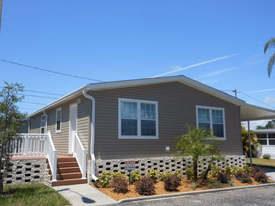 Exterior view of a single-story manufactured home with beige siding, white trim, and a small front porch with stairs. There are two windows visible on the front side, a small palm tree, and other landscaping plants in front of the home. The sky is clear and blue.