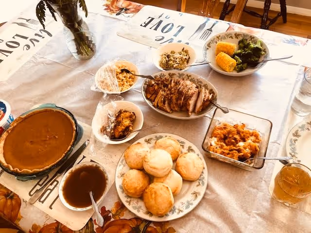 A dining table set with a variety of home-cooked dishes including a pumpkin pie, gravy, biscuits, sliced roasted meat, corn on the cob, broccoli, stuffing, and a casserole. The table is covered with a white tablecloth with autumn-themed decorations and a vase with flowers.