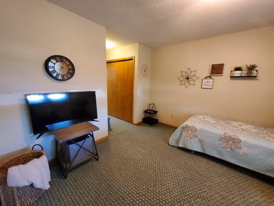 A cozy bedroom with a single bed covered in a floral quilt, a wall-mounted TV on a small wooden stand, a round wall clock above the TV, and decorative wall art including a metal flower, framed quotes, and small potted plants on a shelf. There is a basket with a white towel near the TV stand and a closet with wooden doors in the background.