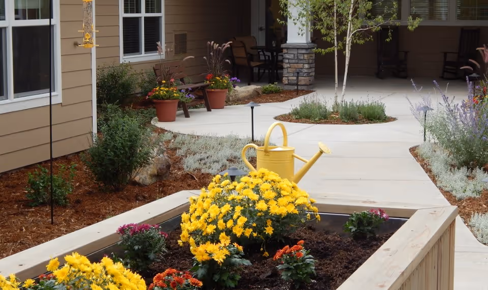 A landscaped outdoor garden area at a senior living facility with a raised wooden flower bed filled with yellow, orange, and purple flowers. A yellow watering can is placed on the edge of the flower bed. The background shows a concrete pathway, potted plants, bushes, and a building exterior with windows and a covered seating area with chairs.