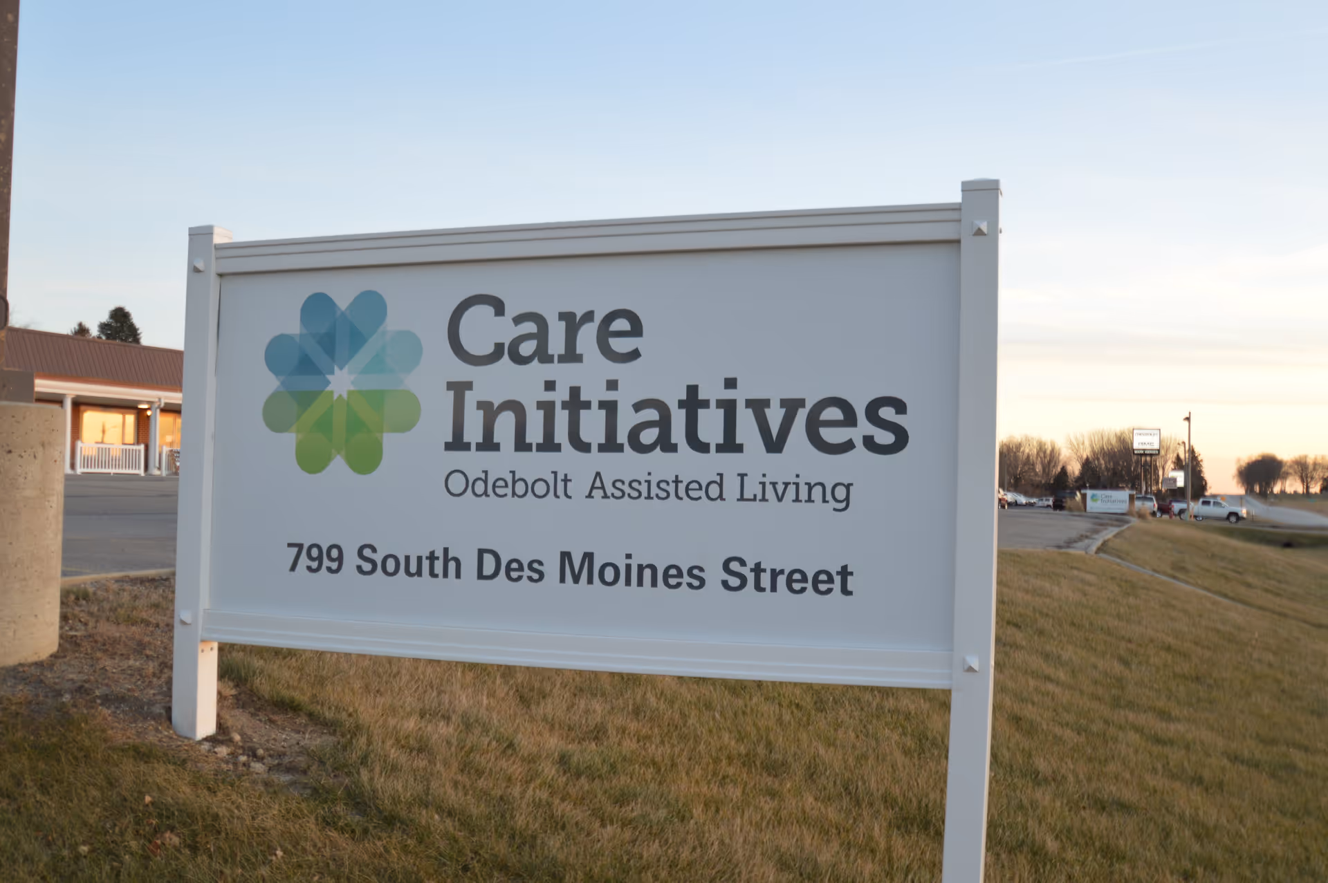 Outdoor sign for Care Initiatives Odebolt Assisted Living located at 799 South Des Moines Street, with a building and parking lot visible in the background during sunset.