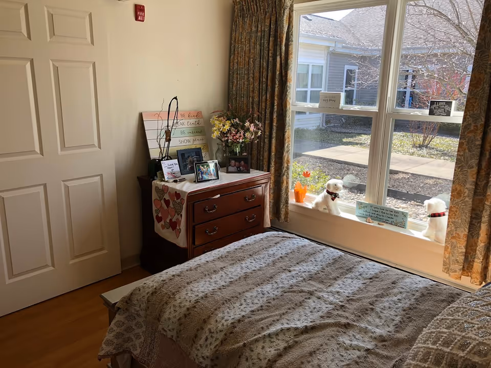 Sunlit bedroom with a bed in the foreground, a wooden dresser topped with framed photos and flowers, and a large window with decorative stuffed animals on the sill.