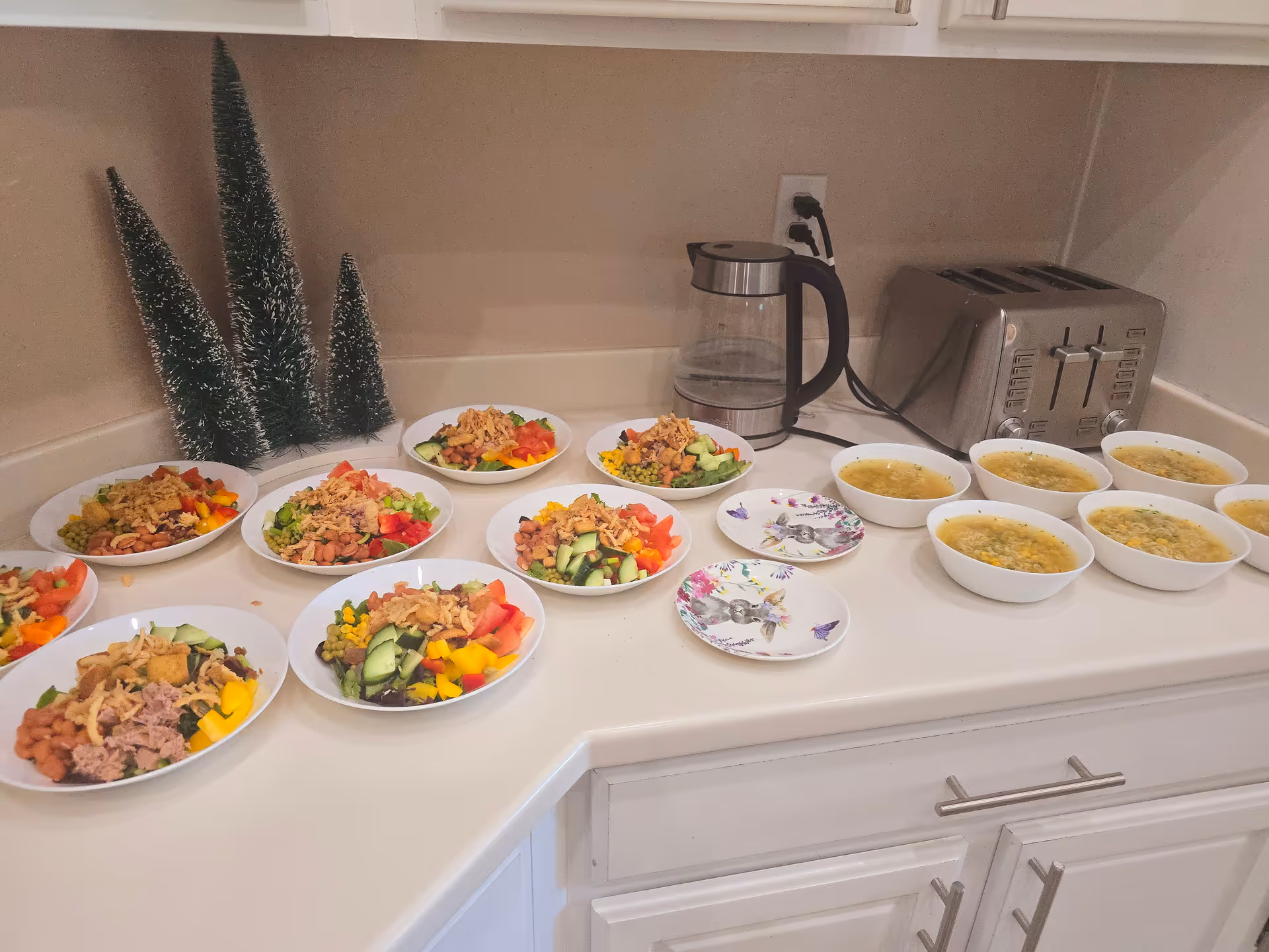 A kitchen countertop with several bowls of salad and soup arranged in rows. The salads contain ingredients like cucumbers, tomatoes, corn, beans, and tuna. There is a glass electric kettle and a stainless steel toaster on the counter. Three small decorative artificial pine trees are placed in the corner.