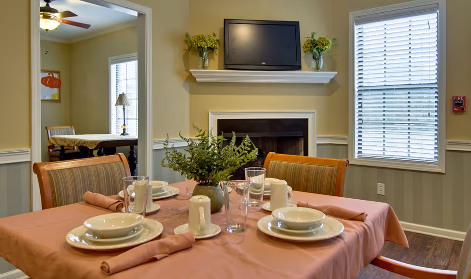 A dining room table set for four with plates, bowls, glasses, mugs, and folded napkins on a pink tablecloth. A green plant centerpiece is in the middle of the table. In the background, there is a fireplace with a TV mounted above it, two vases with flowers on the mantel, a window with blinds, and an adjacent room with another table and chairs.