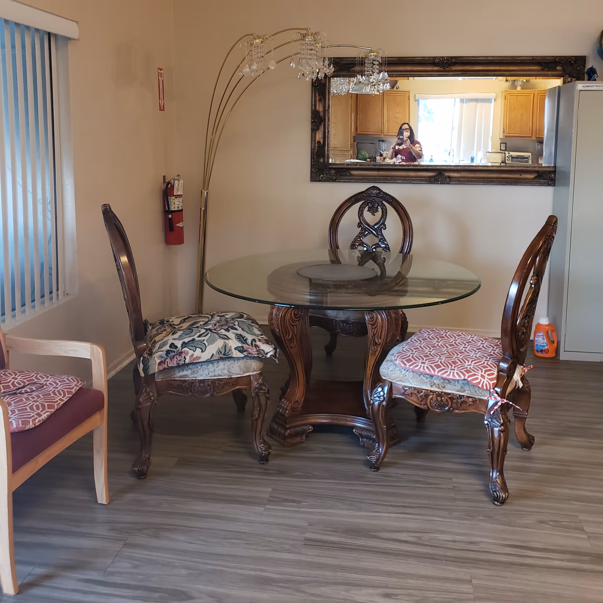 A small dining area with a round glass-top table and three ornate wooden chairs with cushions. A large decorative mirror hangs on the wall reflecting a kitchen area and a person taking the photo. A tall floor lamp with multiple lights is positioned near the table. A fire extinguisher is mounted on the wall, and a window with vertical blinds is visible on the left side.