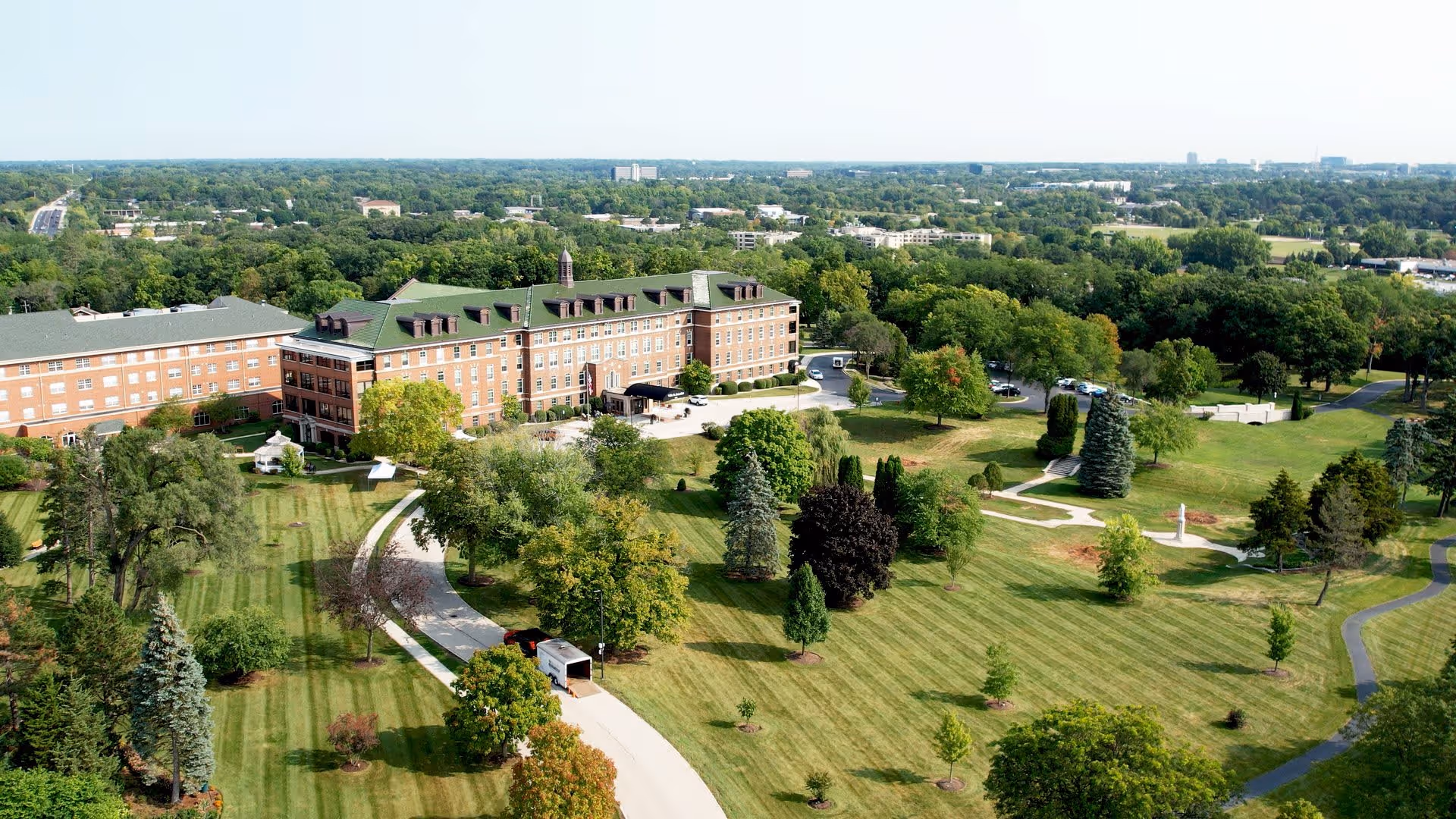 Aerial view of Villa St. Benedict, showing a large brick building with a green roof surrounded by expansive, well-maintained lawns with trees and walking paths.