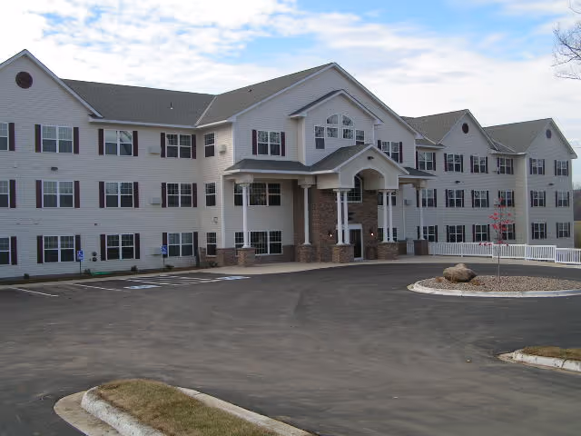 Front exterior view of a large, three-story senior living facility building with white siding, multiple windows, and a covered entrance supported by columns. The paved driveway and parking area are visible in front, along with a small landscaped roundabout with rocks and a young tree.
