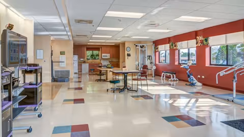 A spacious rehabilitation room with exercise equipment along the right wall, tables and chairs in the center, and a kitchenette area at the far end. The room has large windows letting in natural light and a tiled floor with colored square patterns.