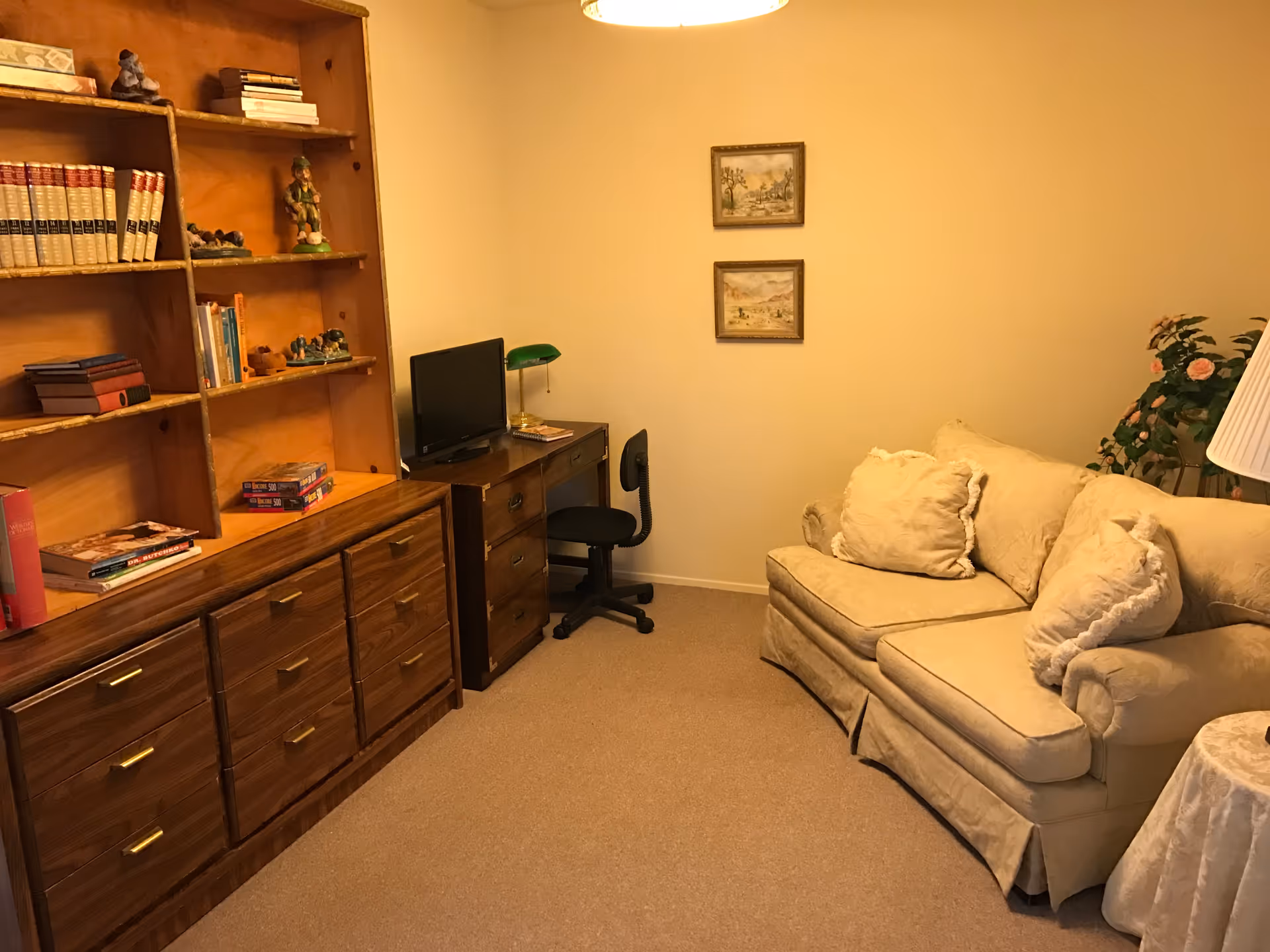 A cozy living room with a beige couch adorned with three cushions, a wooden bookshelf filled with books and decorative items, a small desk with a computer monitor and a green desk lamp, a black office chair, two framed pictures on the wall, a potted plant, and a floor lamp with a white shade.
