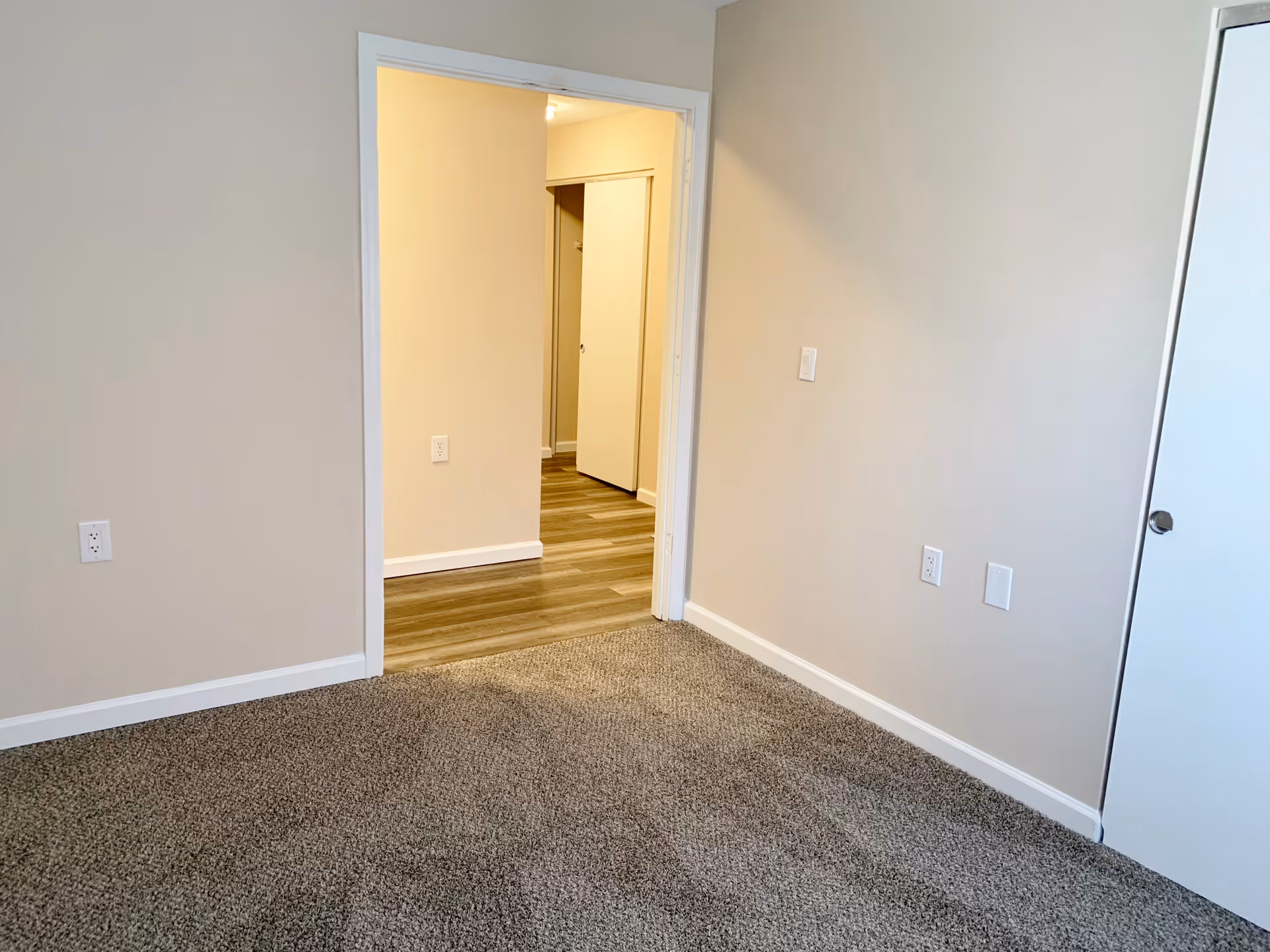 Empty room with beige walls, gray carpeted floor, and a white door leading to a hallway with wood flooring. Several electrical outlets are visible on the walls.