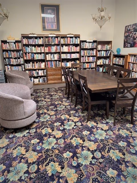 A cozy library room with multiple bookshelves filled with books along the walls. In the center, there is a large wooden table surrounded by eight chairs. Two upholstered armchairs are positioned on the left side. The room has a patterned carpet with floral designs and is decorated with framed pictures and a chandelier hanging from the ceiling.