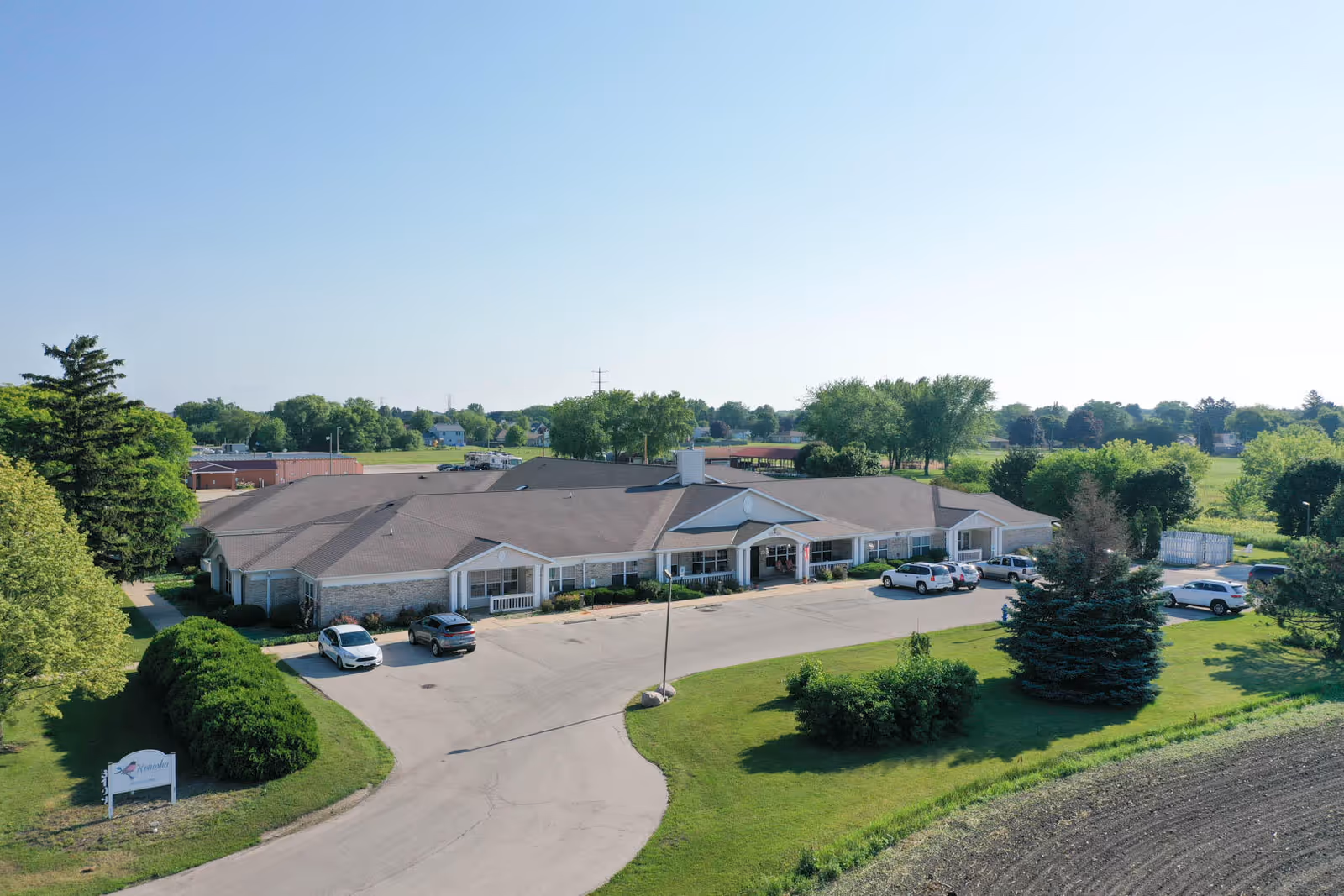Aerial view of a single-story senior living facility building with a large parking area in front, surrounded by green lawns, trees, and open fields under a clear blue sky.