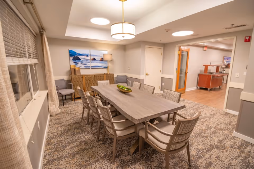 A well-lit dining room with a long wooden table surrounded by upholstered chairs, artwork on the wall, and a service cart visible through an open doorway.