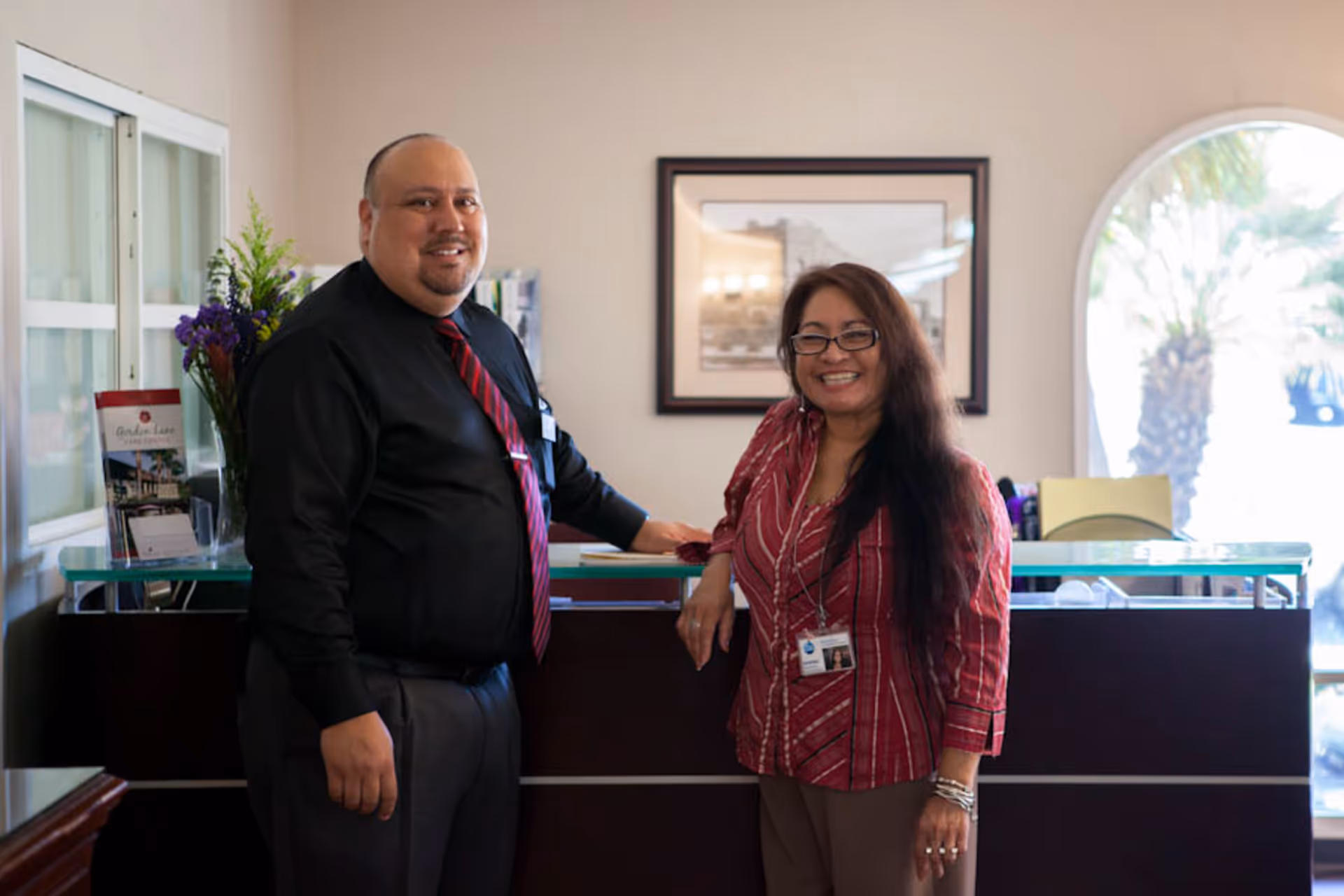 Two smiling staff members standing behind a reception desk inside a care facility. The man on the left is wearing a black shirt with a red and black striped tie, and the woman on the right is wearing glasses and a red patterned blouse with a name badge. Behind them is a framed picture on the wall and a window showing palm trees outside.