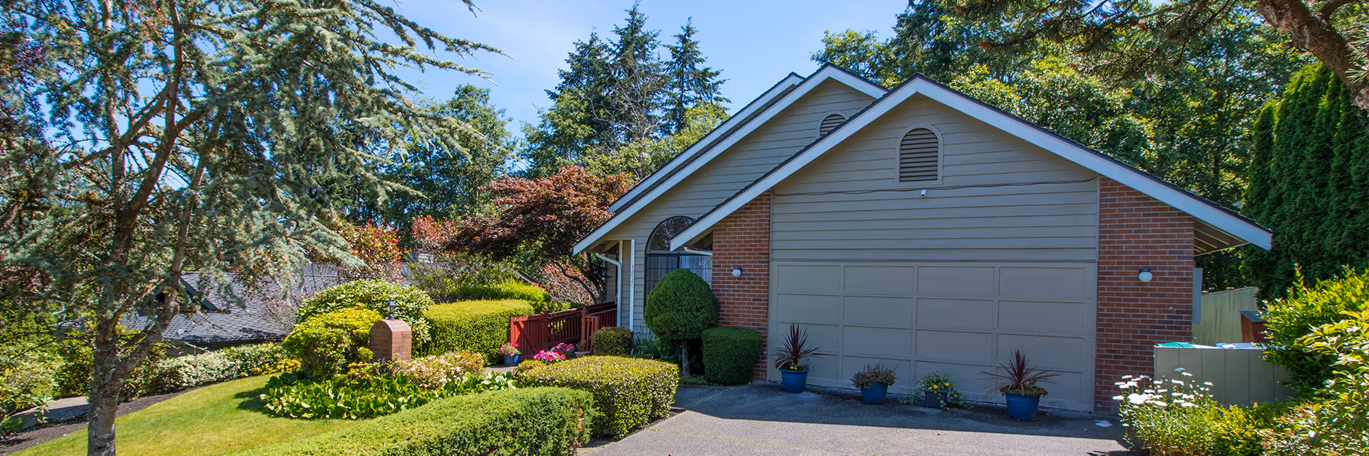 Front exterior of a single-story house with a two-car garage, driveway, and landscaped yard with shrubs and trees.