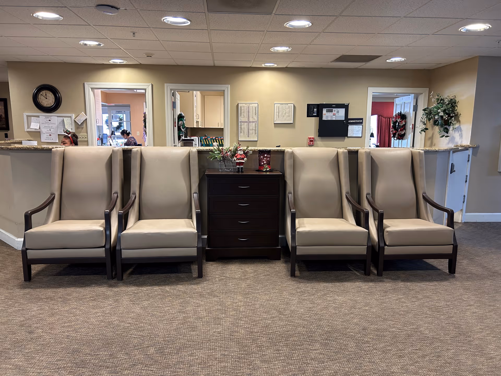 A waiting area with four beige armchairs arranged in two pairs on either side of a dark wooden cabinet with drawers. The cabinet has holiday decorations on top, including a small Santa figurine and a candle holder. Behind the chairs is a reception desk with two open doorways and a window, showing staff working inside. The walls are beige, and the floor is carpeted.