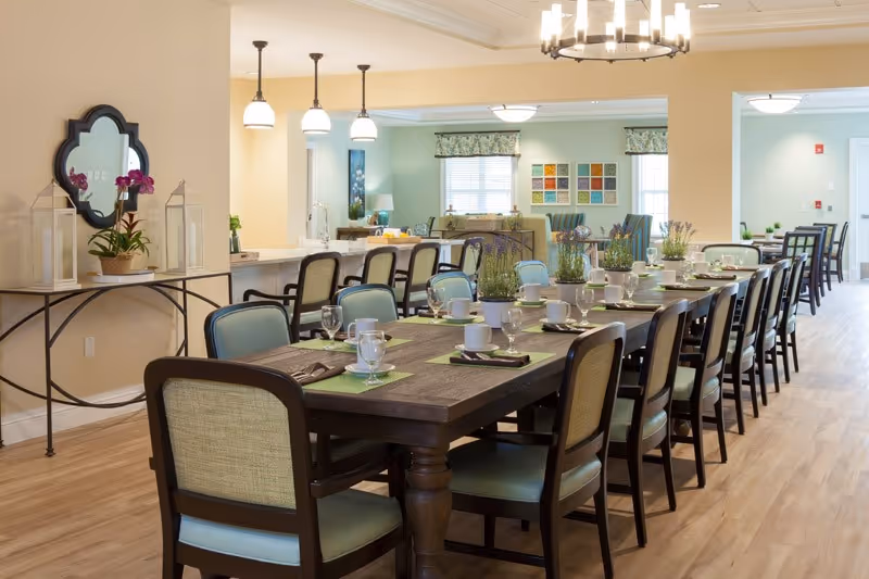 A spacious dining room with a long wooden table set for a meal, featuring place settings with glasses, cups, and napkins. The room has light-colored walls, wooden flooring, and several chairs around the table. In the background, there is a kitchen area with bar stools and pendant lights, and a living area with windows and colorful wall art.