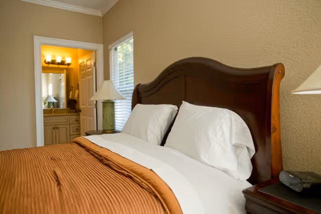 A neatly made bedroom with a wooden headboard, bedside lamps, and a view into an adjoining bathroom.