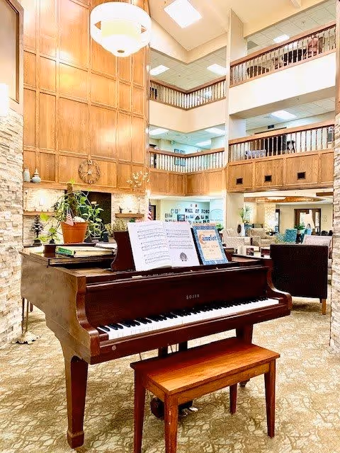 Interior view of a senior living facility common area featuring a grand piano with sheet music and a plant on top. The space has high ceilings with wooden paneling and railings on the upper floors. Comfortable seating areas with armchairs are visible in the background, along with warm lighting and a cozy atmosphere.