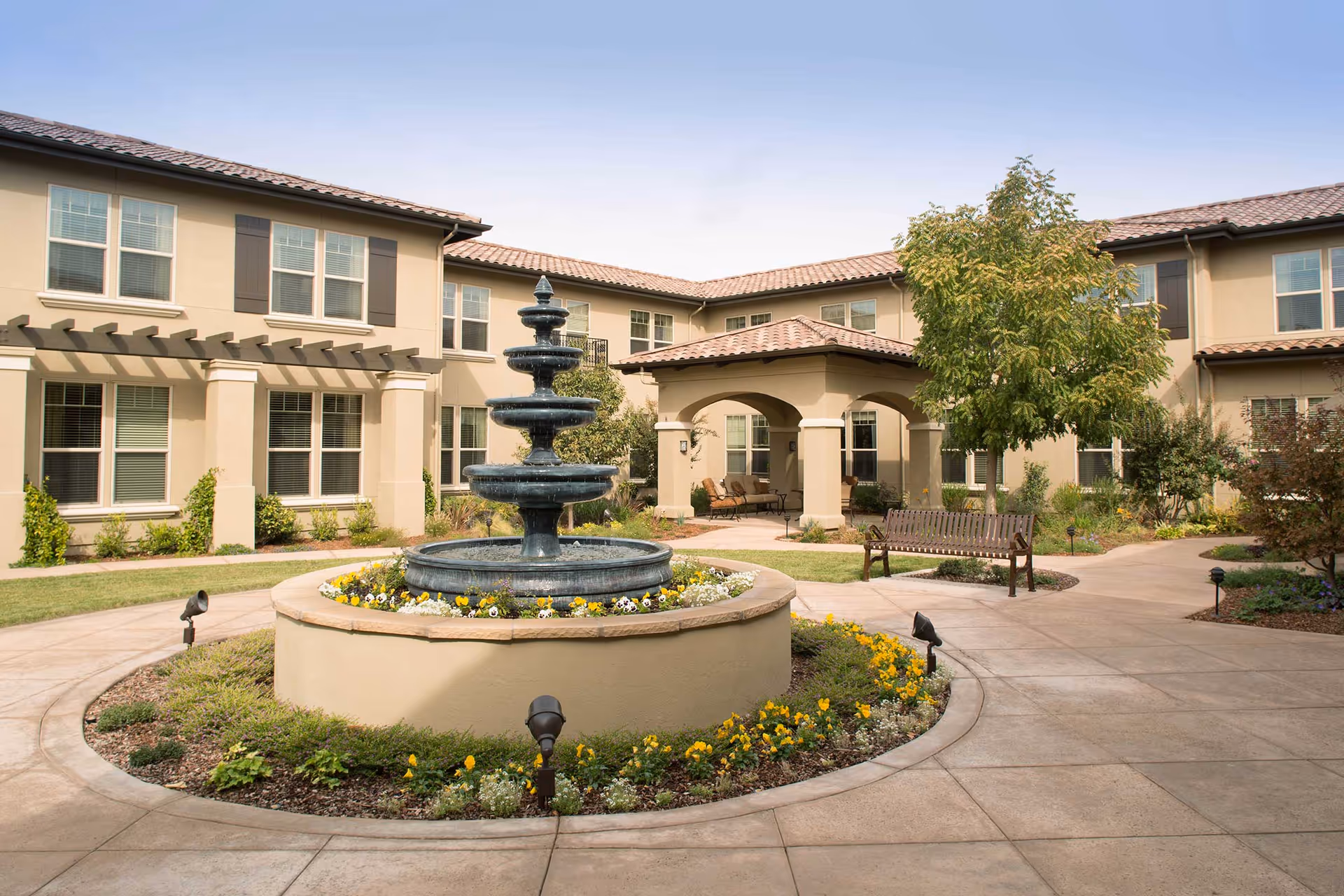 Courtyard with a tiered fountain, flower beds and benches in front of a two-story residential building.