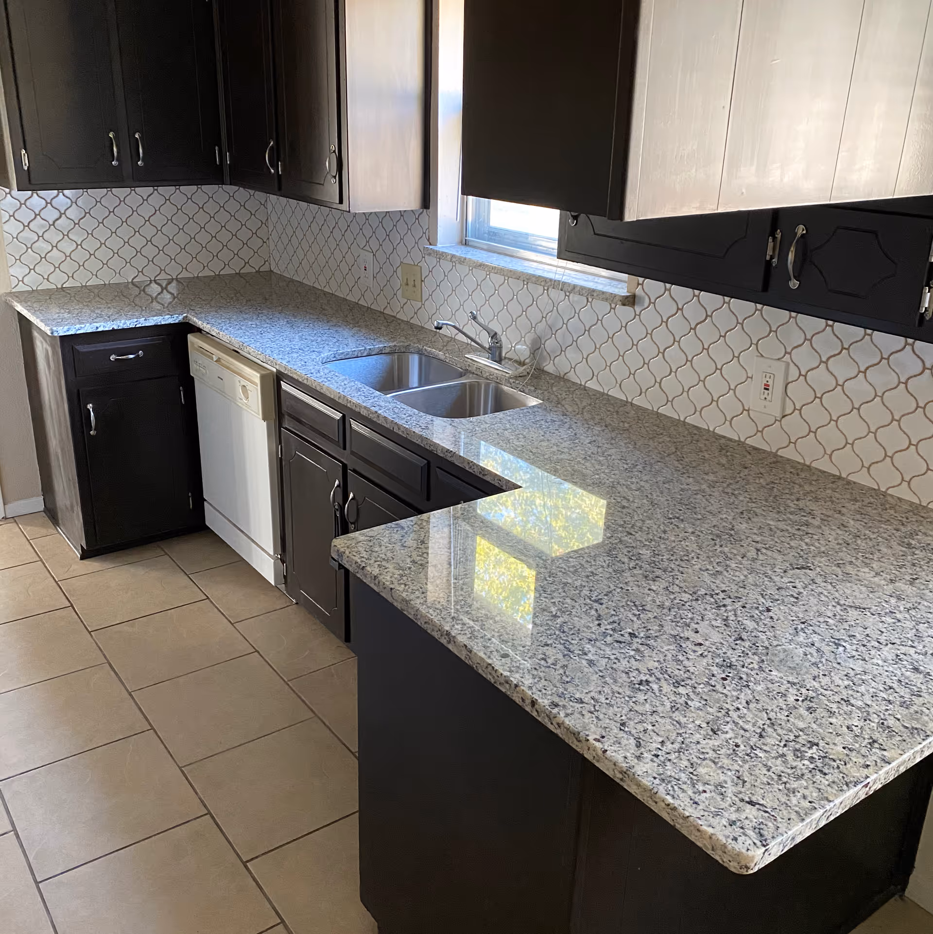 A kitchen interior featuring dark wooden cabinets, a granite countertop with a double stainless steel sink, a white dishwasher, and a tiled backsplash with a geometric pattern. There is a window above the sink allowing natural light into the space.