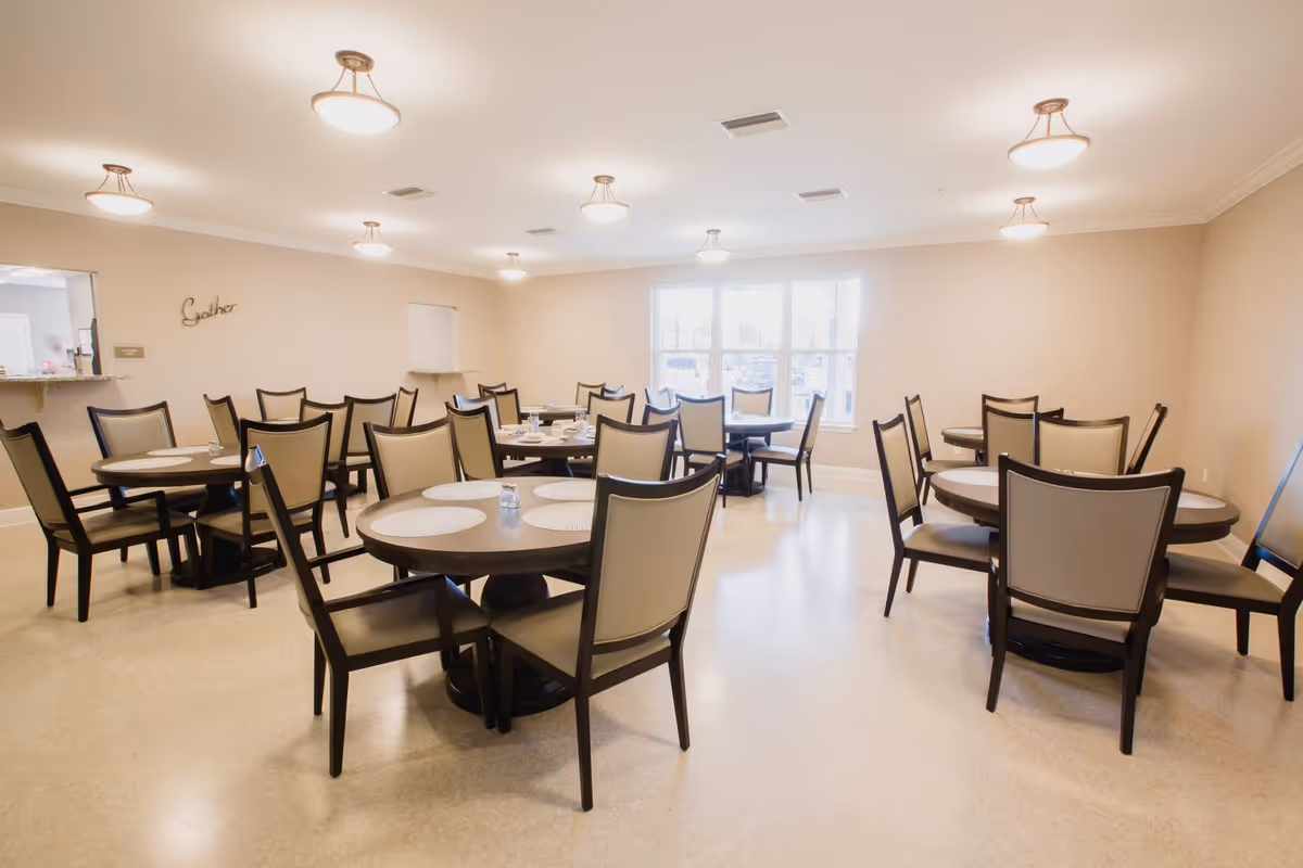 A bright dining room with several round tables and beige cushioned chairs arranged neatly. The room has beige walls, multiple ceiling lights, and a large window letting in natural light. A small serving window is visible on one wall with the word 'Gather' displayed above it.